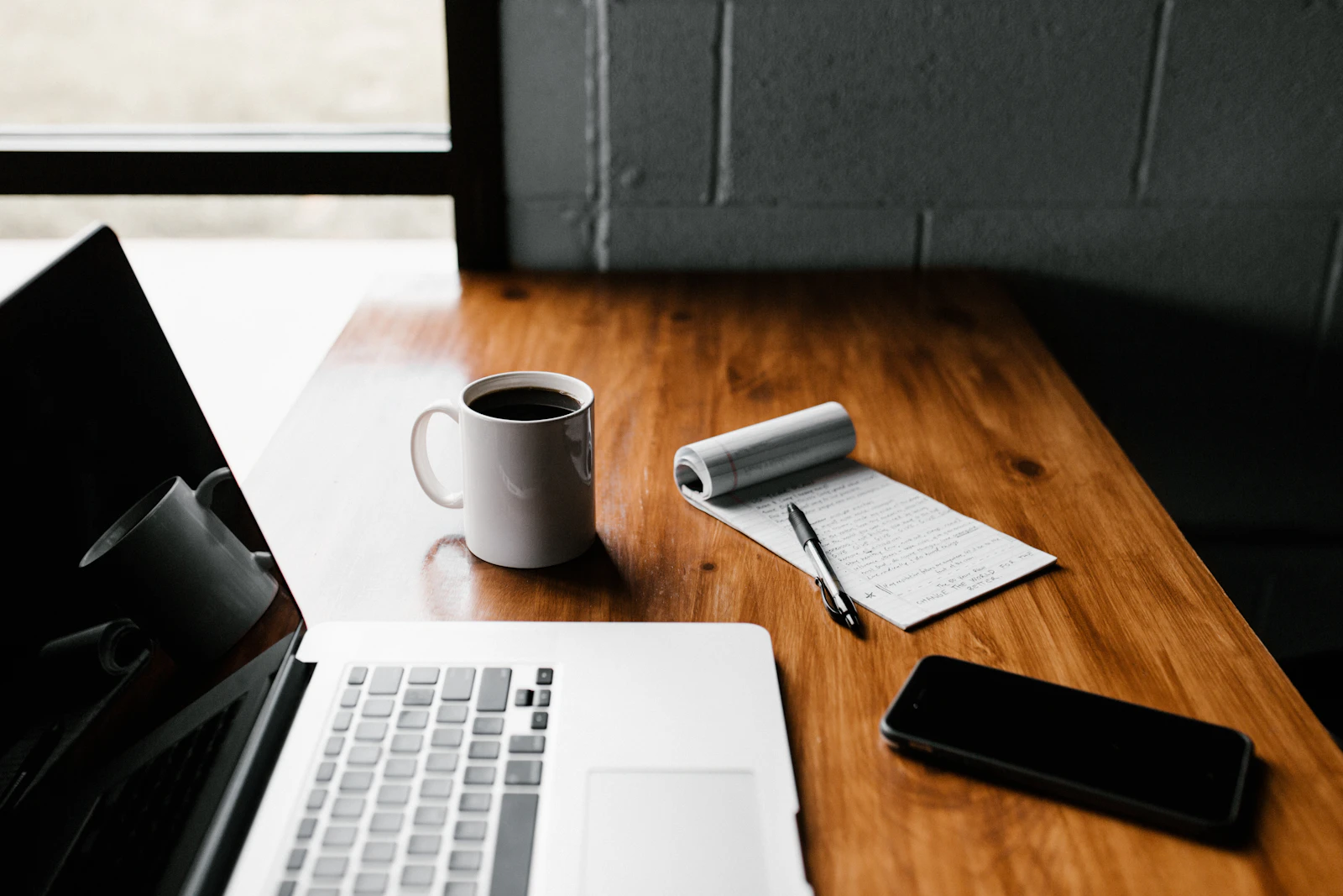A laptop, coffee mug, notepad and phone on a wooden desk