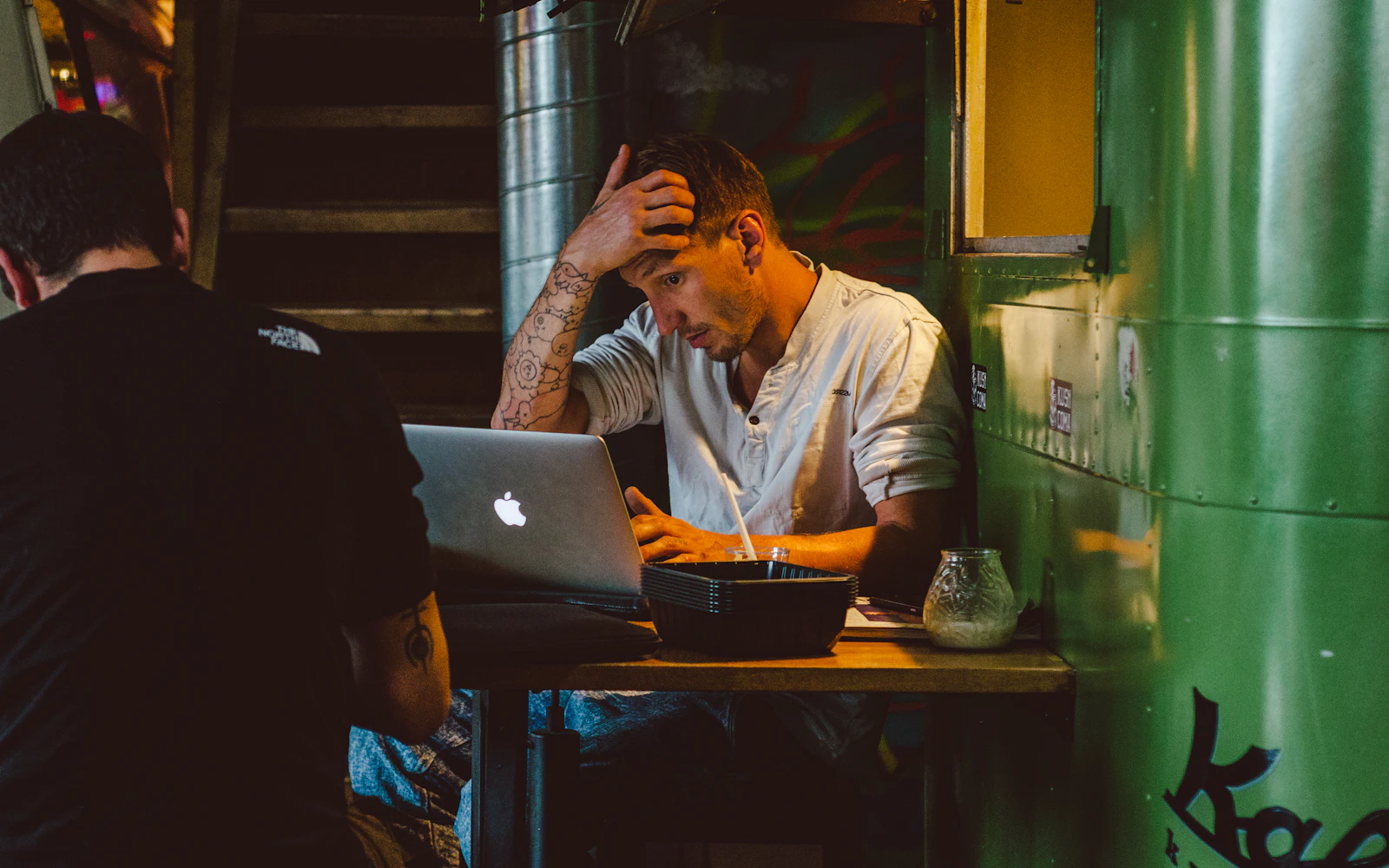 A man at a laptop in a cafe, hand on his forehead, waiting for a page to load